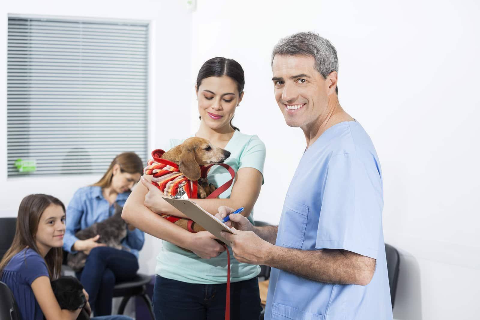 Male nurse in blue scrubs writes on clipboard while woman holds dachshund in waiting room.